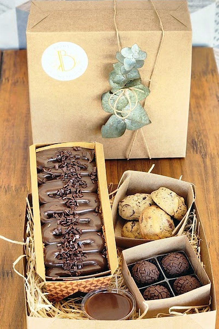 Caixa Afeto da Brada, aberta, com bolo de rolo e biscoitos, sobre papel kraft com eucalipto